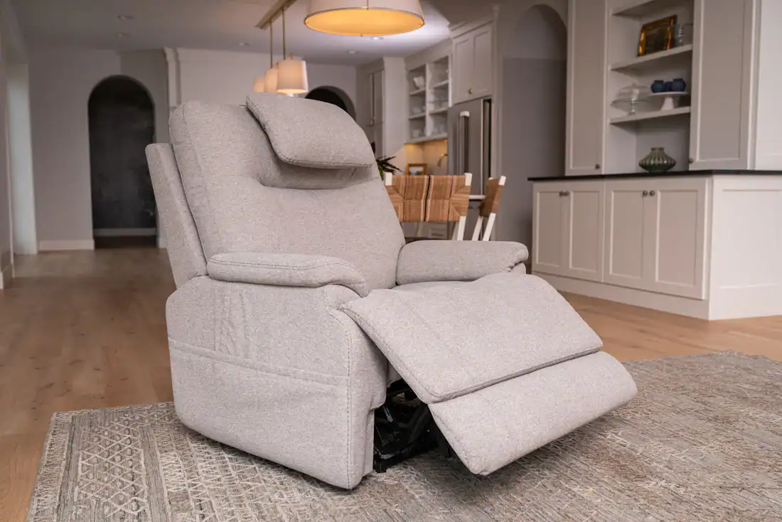A light gray recliner chair with footrest extended sits on a patterned rug in a modern, bright kitchen and dining area with white cabinets and wooden floors.