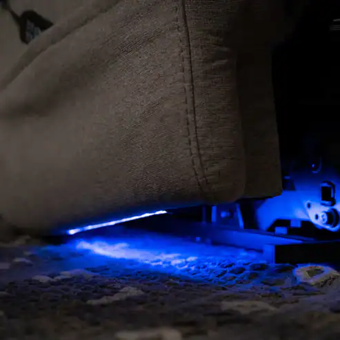 A close-up of the bottom edge of the Model 2 Plus Shell Exclusive grey recliner, featuring blue LED lights that illuminate the area underneath and cast a glow on the patterned carpet.