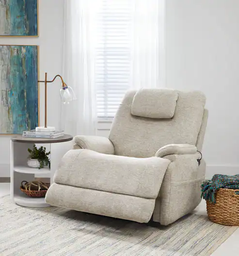 A beige Model 2 Plus Shell Exclusive recliner with plush headrest and footrest sits by a round white side table with a lamp and plant. A woven basket and Shell Exclusive abstract wall art complete the cozy, sunlit room.