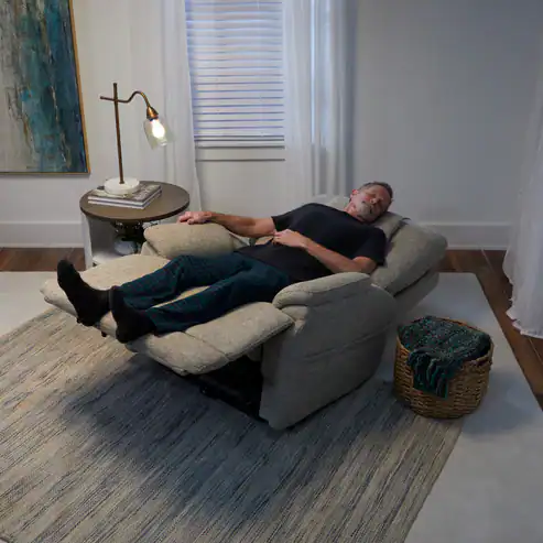 An older man in dark clothing relaxes on a fully reclined beige Model 2 Plus Shell Exclusive armchair with his feet up, positioned on a rug in a cozy, softly lit living room beside a table, lamp, and basket.