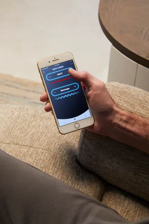 Sitting on a beige couch near a round wooden table, a person uses their smartphone to control the heat and massage features of the Model 2 Plus Shell Exclusive recliner—demonstrating its comfort and convenience.