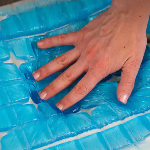 A hand presses down on a surface covered with Model 2 Plus Shell Exclusive blue gel packs, making the gel shift and indent under the pressure.