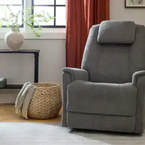 A Zecliner Model 3+ Umber Power Sleep Chair sits on a light rug near a woven basket with a blanket, a console table with a vase, and a window with red curtains in a cozy, well-lit room.