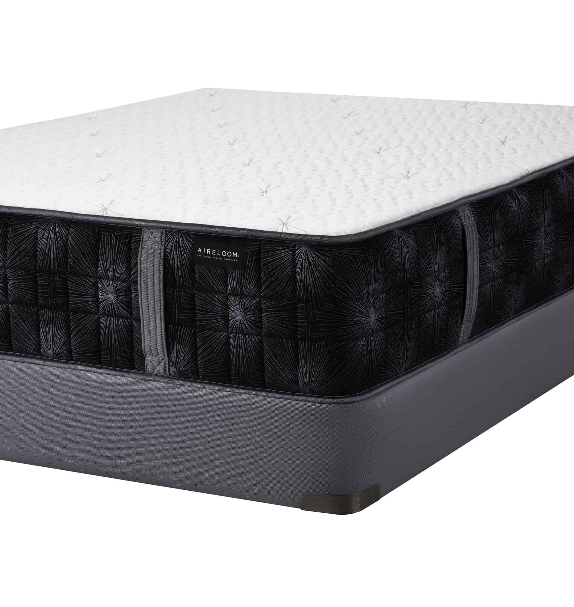 A close-up view of the Summit Luxury Firm mattress shows its white quilted top, black patterned sides, gray base, and a visible Aireloom tag, emphasizing its firm support and Summit-inspired luxury design.