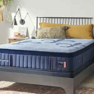 A modern bedroom with a neatly made Stearns & Foster Lux Hybrid Elite 14.5″ Medium bed, mustard and green pillows, black metal headboard, wall lamp, decorative accents, and a plant adding warmth to the space.