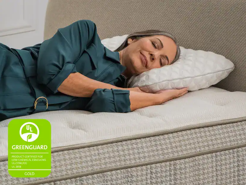 A woman with gray hair lies on her side, smiling with her eyes closed on a bed with a white pillow and the Leesa Reserve Hybrid Mattress, featuring a UL GREENGUARD Gold certification label in the lower left corner.