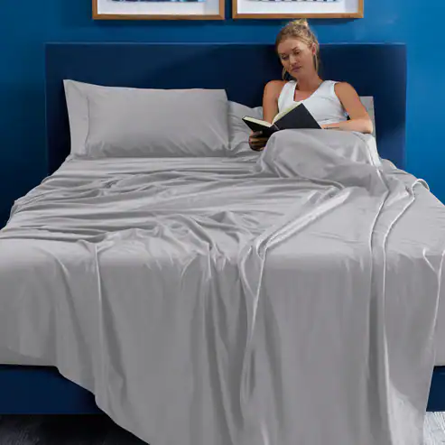 A woman sits upright in bed reading a book. The bed is covered with Hyper - Cotton Performance Sheets and light gray pillowcases, and has a blue headboard. Two framed pictures hang on the blue wall behind her.
