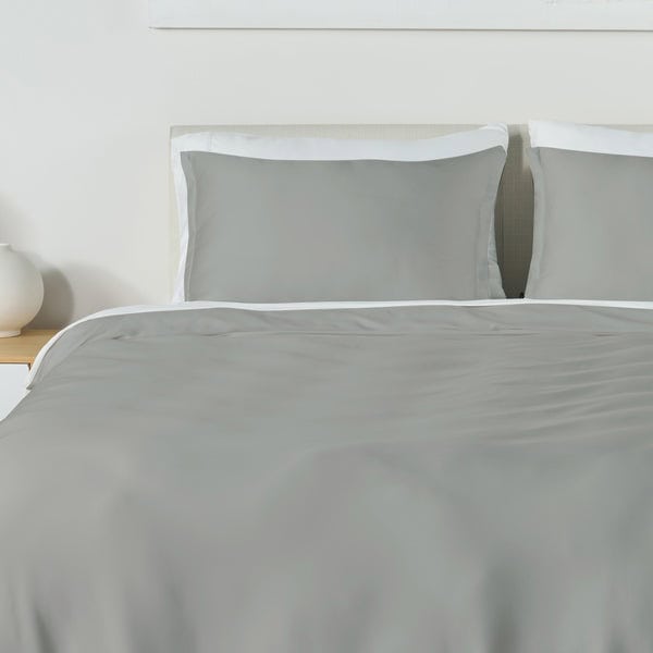 A neatly made bed with the Performance Duvet & Sham Set - Solid in gray, featuring shams bordered in white trim, sits against a light headboard and wall. On the left, a white nightstand holds a round white lamp.