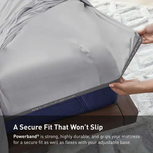 A person tucks a fitted gray Hyper-Cotton Performance Sheet with a labeled Powerband® elastic edge over a blue mattress on a wooden bed frame; text highlights the secure, non-slip fit.