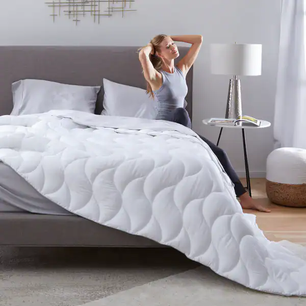 A woman stretches on the edge of a neatly made bed with a white Performance Comforter—available in light, medium, and ultra weights—in a bright, modern bedroom.
