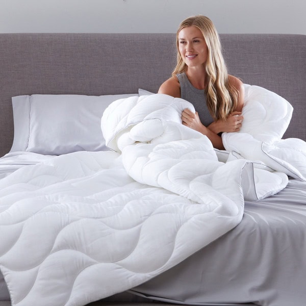 A woman with long blonde hair smiles while sitting on a bed with light gray sheets and a gray headboard, wrapped in the medium weight Performance Comforter.