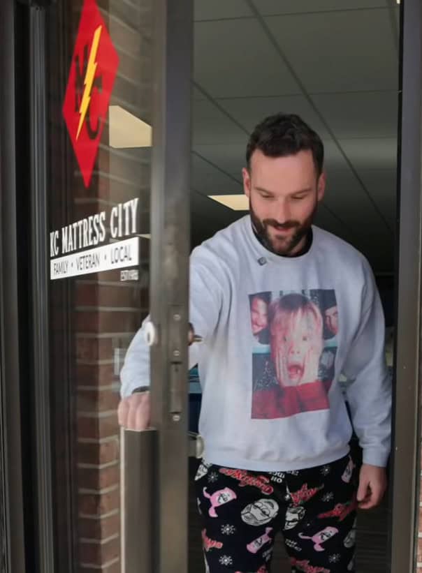 A man wearing a "Home Alone" sweatshirt and festive pajama pants opens the glass door of KC Mattress City, a shop with a sign reading "Family • Veteran • Local," ready to chat about Christmas plans.