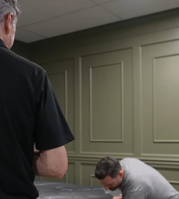 Two men in a room with green paneled walls; one stands in the foreground with his back to the camera, while the other bends over a table—perhaps discussing a company's promise or plan.