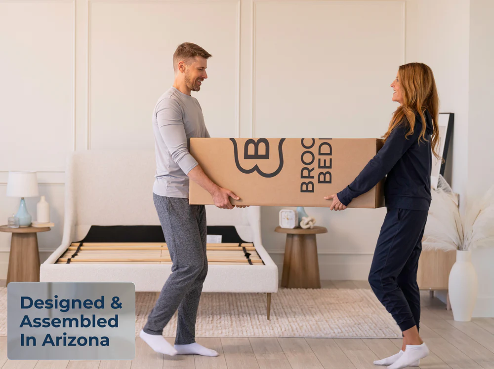 A smiling couple in pajamas carries a ThermoBalance Soft mattress box into a sunlit bedroom with an unassembled bed frame. A corner sign says, "Designed & Assembled in Arizona.