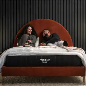 A smiling couple lounges on a bed with "TITAN+ CORE" on the side, showcasing the Brooklyn Titan Plus Mattress (Copy). The rust-colored frame stands between two black nightstands with modern lamps, set against a ribbed black wall.
