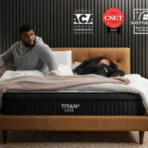 A man sits up while a woman lies down on a large bed in a modern bedroom. The bed, labeled "Brooklyn Titan Plus Core Mattress," is showcased with award logos for Brooklyn Titan Plus above the headboard.