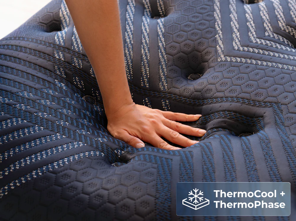 A hand presses on a dark, textured mattress with blue patterns. Icons and text in the corner read "ThermoCool + ThermoPhase," featuring snowflake and phase symbols to highlight the ThermoBalance Elite comfort system.