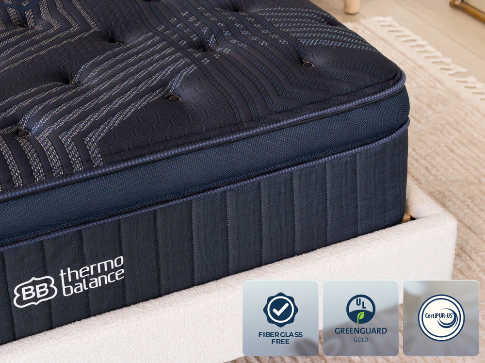 Close-up of the ThermoBalance Elite dark blue mattress on a beige bed frame, featuring "BB thermo balance" label and badges for Fiberglass Free, GREENGUARD Gold, and CertiPUR-US certifications at the bottom.