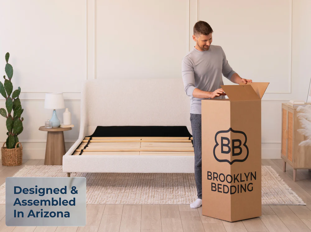 A man opens a large Brooklyn Bedding box featuring ThermoBalance Elite in a bright bedroom with an unassembled bed frame. Nearby, a side table holds a lamp and vase. Text displays: "Designed & Assembled In Arizona.