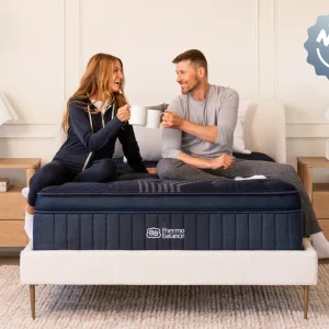A smiling couple sits on a bed with the ThermoBalance Elite mattress, holding white mugs in a cozy modern bedroom. A "NEW" badge is displayed in the top right corner.