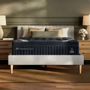 A neatly made bed featuring the DreamCloud Luxe Hybrid Mattress sits between two wooden nightstands with lamps, complemented by decorative pillows and a bouquet of white flowers in a cozy, modern bedroom.