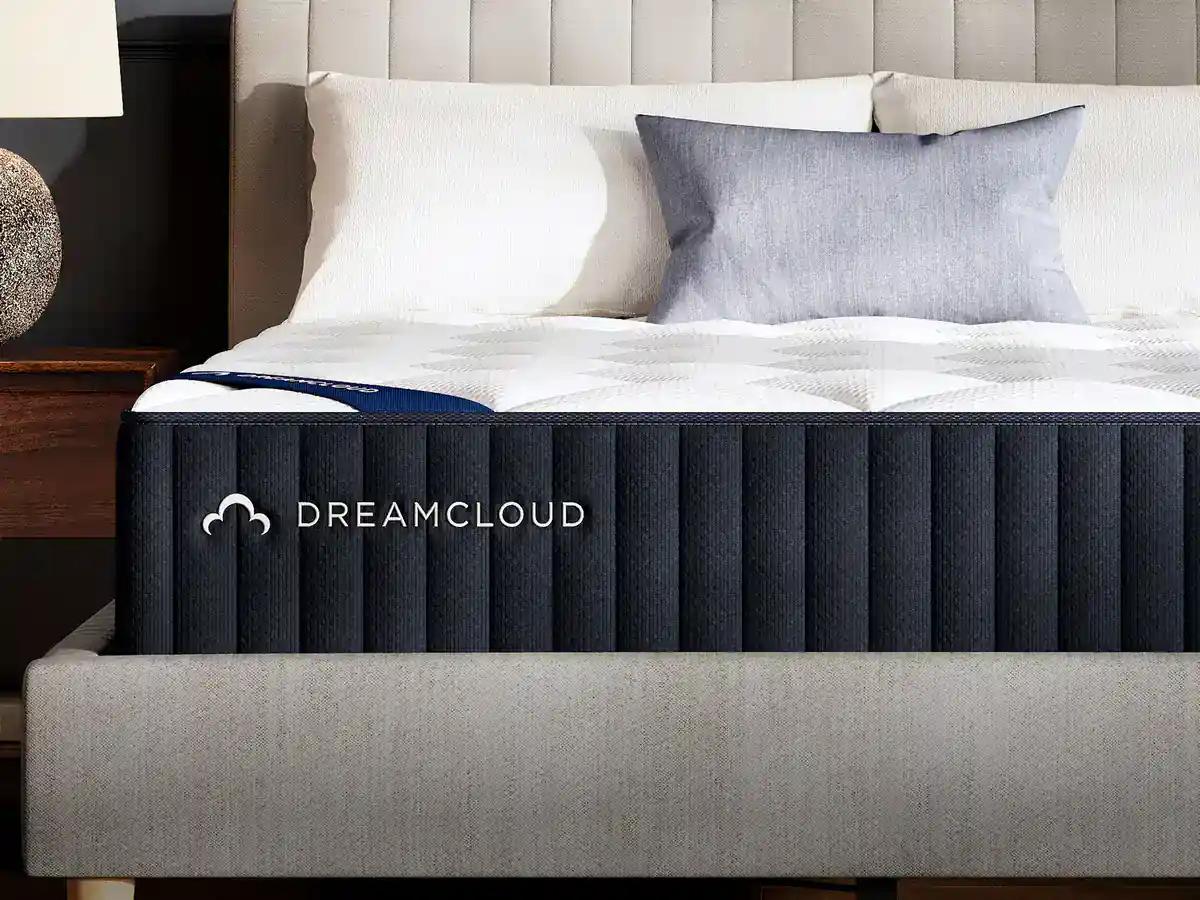 A close-up of a DREAMCLOUD mattress on a beige upholstered bed frame, with a grey and blue pillow resting against the headboard; a wooden nightstand is partially visible on the left.