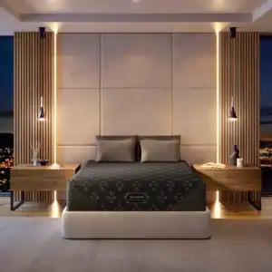 A modern bedroom with a large upholstered headboard, dark geometric bedding on a Beautyrest Black® Series One 14.25" Medium Mattress, wooden nightstands, pendant lights, and floor-to-ceiling windows overlooking the city.