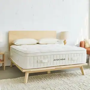 A modern bedroom featuring a light wood bed frame, a white Avocado Luxury Organic Mattress – Plush Organic Latex Hybrid Mattress labeled “Avocado,” two white pillows, two bedside tables with lamps, an orange chair, and a white textured rug on a concrete floor creates the perfect setting for those who appreciate the comfort of a luxury organic mattress.