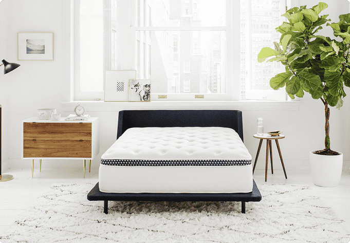 A modern bedroom showcases The WinkBed Luxury Hybrid Mattress &ndash; 4 Firmness Options set on a dark bed frame, complemented by a white rug, wooden nightstand, potted plant, and large windows that fill the space with natural light. With its multiple firmness choices, this room blends comfort and style for the ultimate retreat.